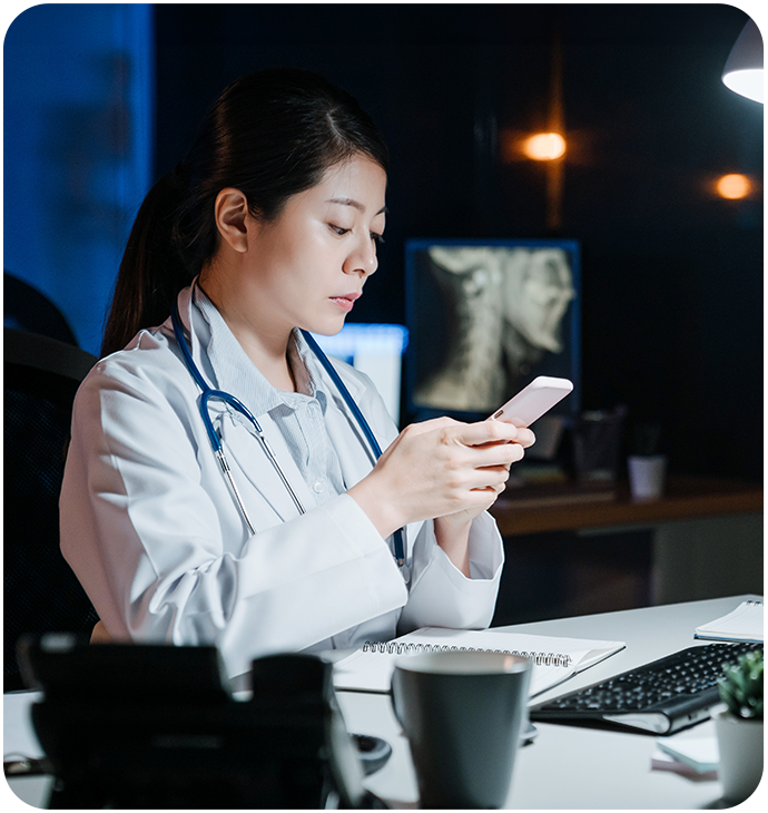 A female doctor in a white coat, with a stethoscope around her neck, sitting at a desk in a dimly lit office. She is focused on her smartphone, with a computer monitor and X-ray images visible in the background. The desk is cluttered with a keyboard, notepad, and coffee cup